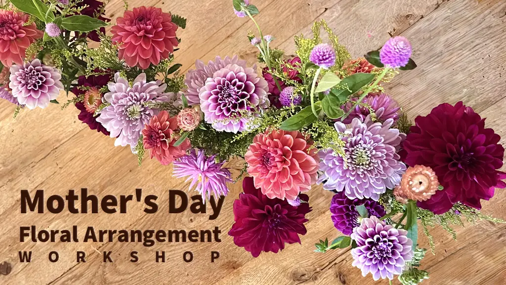 A decorative graphic for a Mother's Day Floral Arrangement Workshop, set against a light-brown textured wood-grain background. Large, calligraphic and serif text on the left reads, 'Mother's Day' in a large, elegant script with decorative flourishes, above 'Floral Arrangement' in a smaller simple serif font, and 'WORKSHOP' in large, bold capital letters at the bottom. A large pink and purple dahlia flower with leaves is positioned in the upper-left corner. The right side of the graphic is bordered by a lush and varied floral arrangement of colorful flowers, thistles, roses, and foliage. Smaller illustrative elements and floral motifs are scattered across the composition, creating a celebratory and vintage feel. The overall presentation is elegant and celebratory.